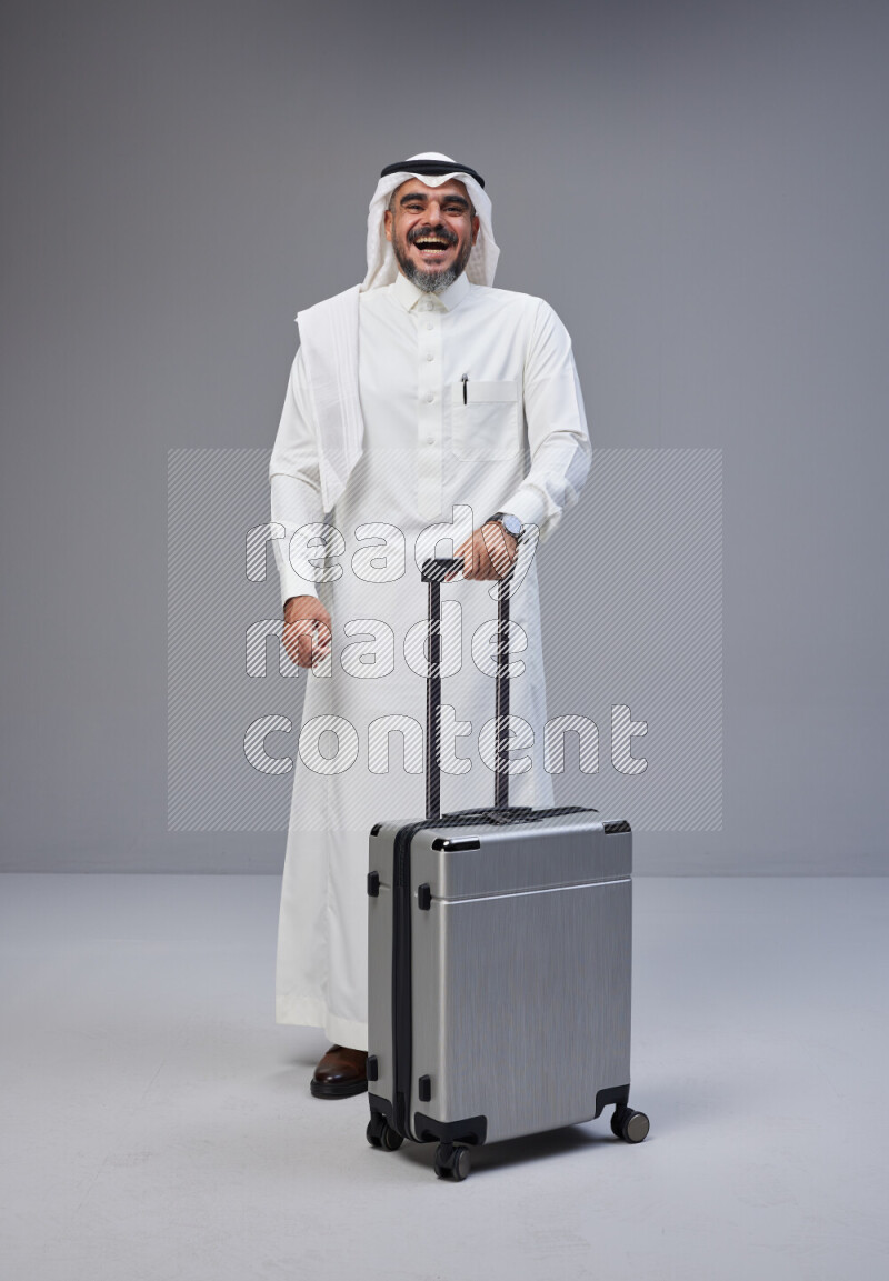 Saudi man wearing Thob and white Shomag standing holding Travel bag on Gray background