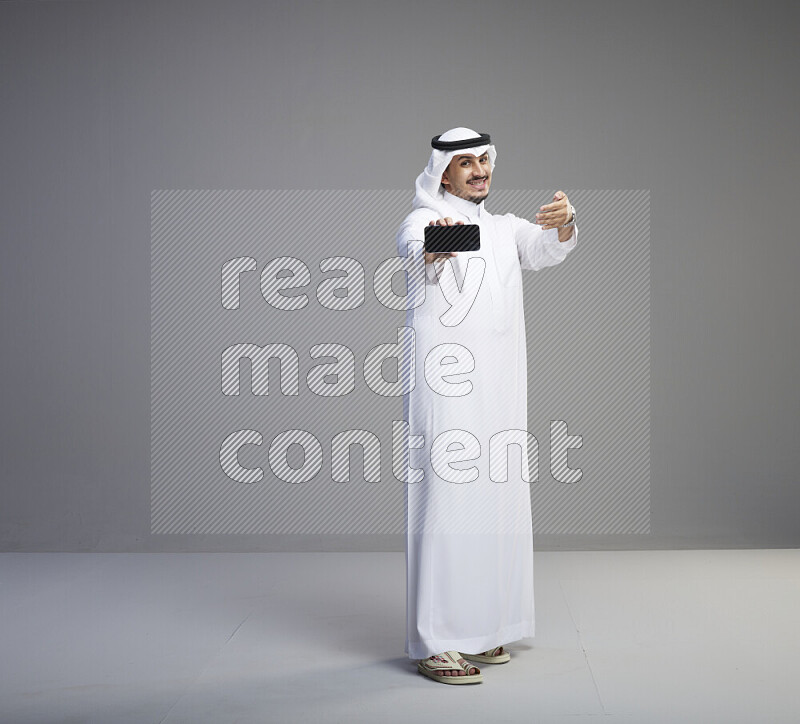 A Saudi man standing wearing thob and white shomag showing phone to camera on gray background