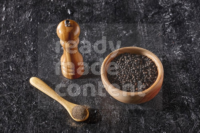 A wooden bowl full of black pepper and a wooden spoon full of black pepper powder and a wooden grinder on a textured black flooring