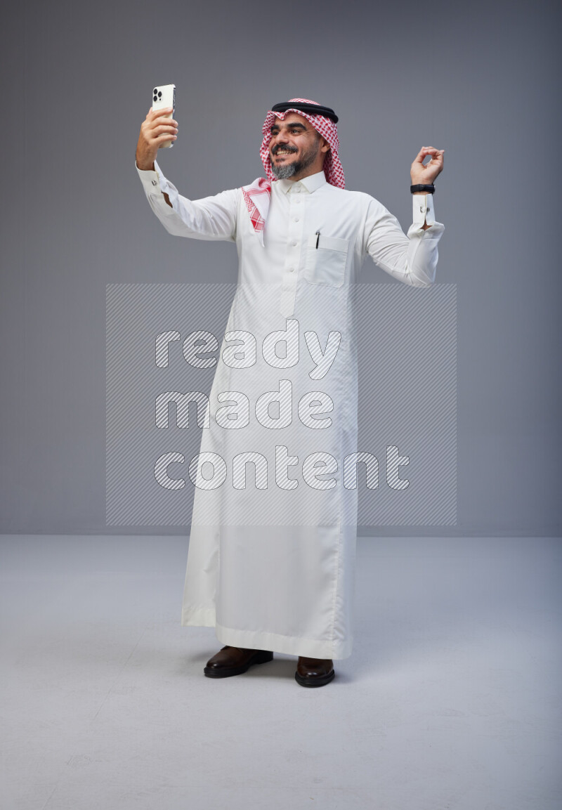 Saudi man Wearing Thob and red Shomag standing taking selfie on Gray background