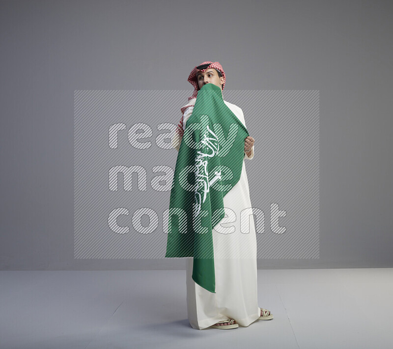 A saudi man standing wearing thob and red shomag holding big saudi flag on gray background