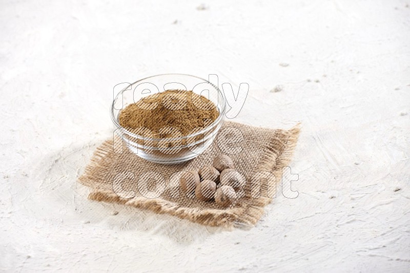 A glass bowl full of nutmeg powder with whole seeds beside it on burlap fabric on a textured white flooring