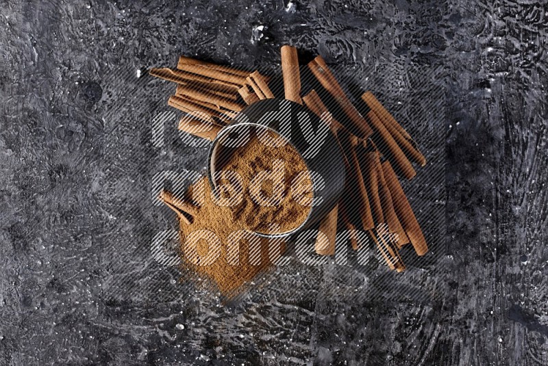 Black pottery bowl over filled with cinnamon powder and cinnamon sticks around the bowl on a textured black background