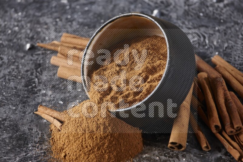 Black pottery bowl over filled with cinnamon powder and cinnamon sticks around the bowl on a textured black background