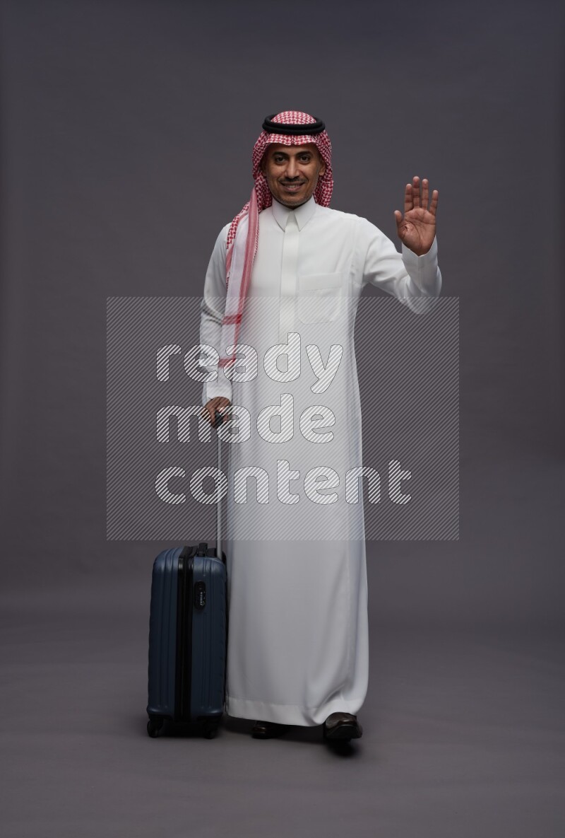 Saudi man wearing thob and shomag standing holding bag on gray background