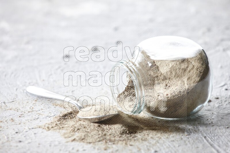 A flipped herbal glass jar and metal spoon full of white pepper powder with spilled powder on textured white flooring