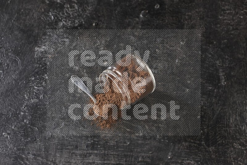 وعاء زجاجي مقلوب وملعقة معدنية ممتلئان ببودرة القرنفل مع حبوب القرنفل المتناثرة على أرضية سوداء