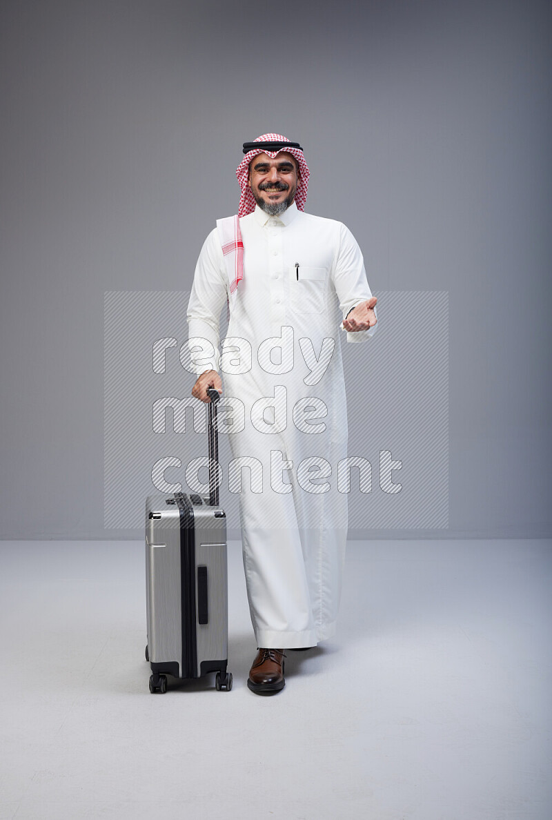 Saudi man wearing Thob and red Shomag standing holding Travel bag on Gray background