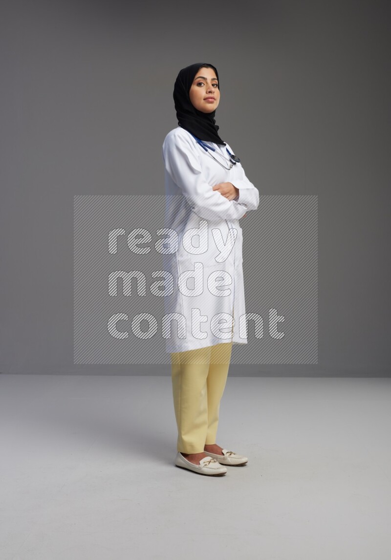 Saudi woman wearing lab coat with stethoscope standing with crossed arms on Gray background