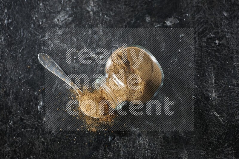 A flipped glass spice jar and a metal spoon full of cumin powder and powder spilled out on a textured black flooring