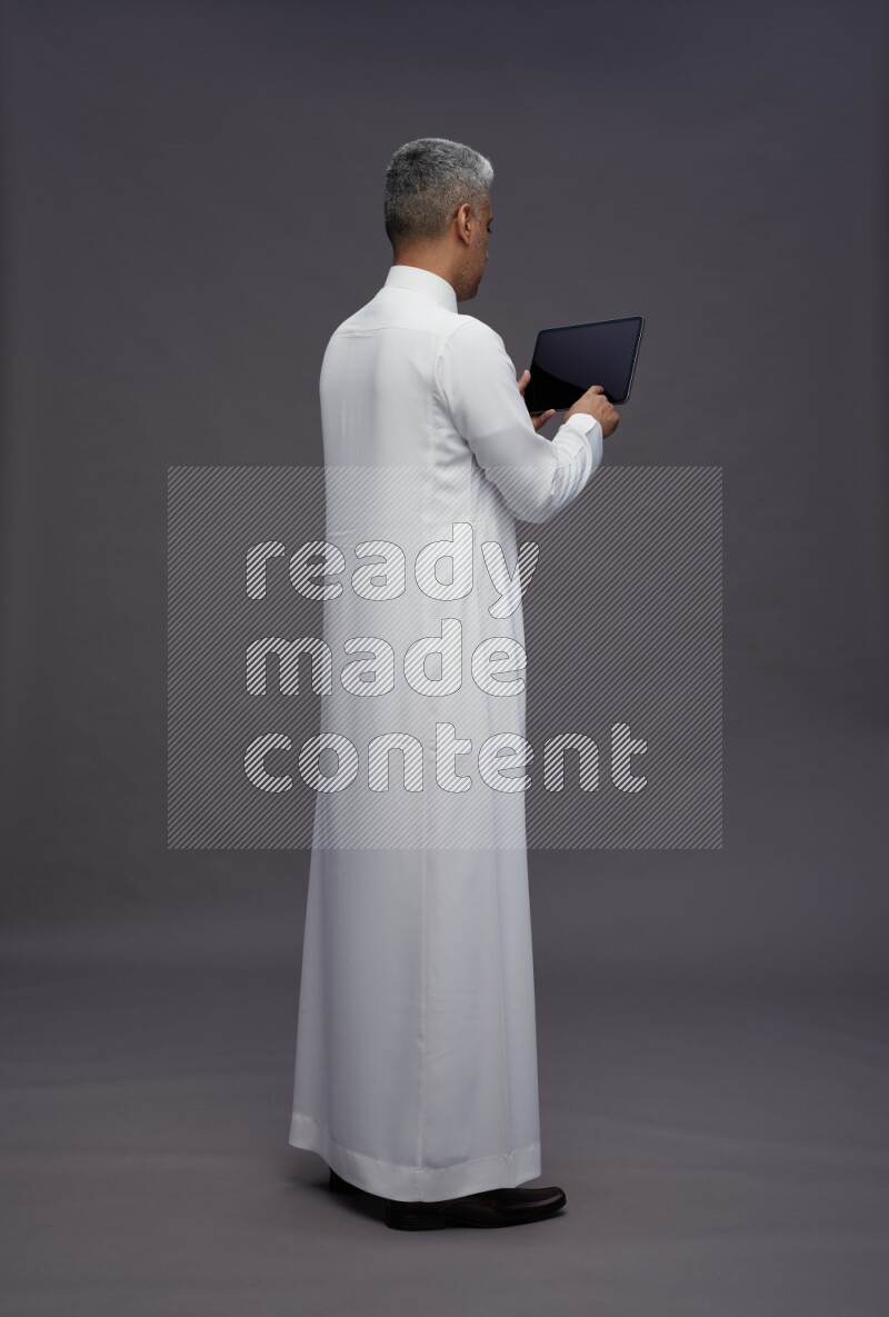Saudi man wearing thob standing working on tablet on gray background