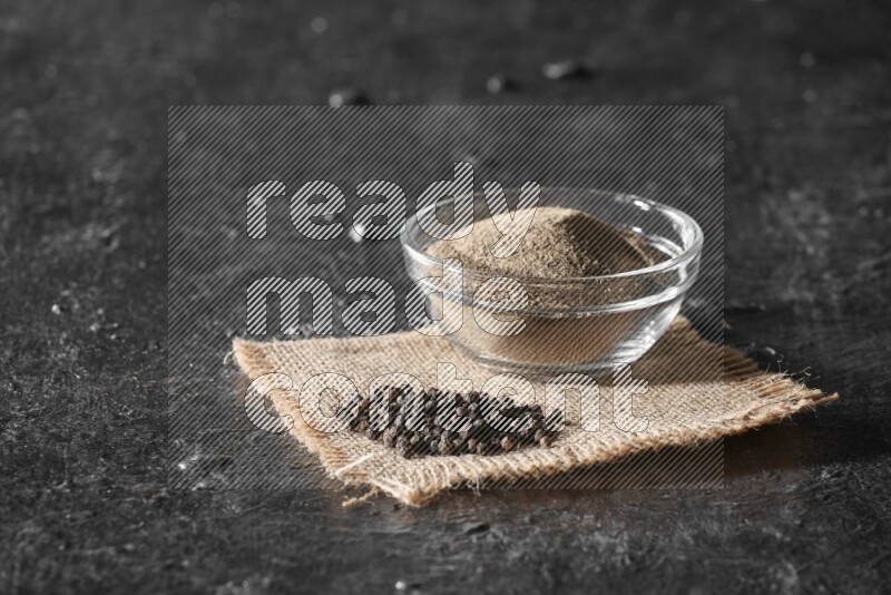 A glass bowl full of black pepper powder and black pepper beads on burlap fabric on textured black flooring
