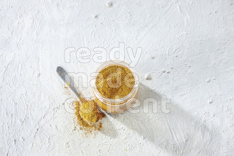 A glass jar and a metal spoon full of turmeric powder on a textured white flooring