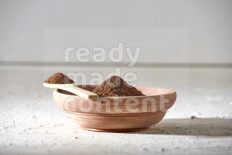 A wooden bowl and a wooden spoon full of cloves powder on a textured white flooring
