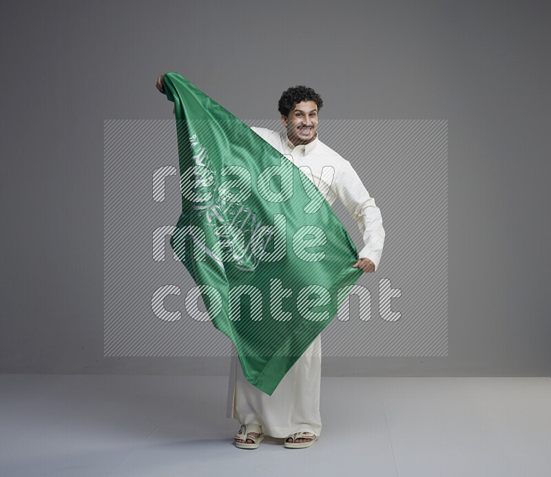 A Saudi man standing wearing thob holding big Saudi flag on gray background