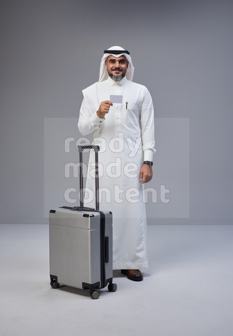 Saudi man wearing Thob and white Shomag standing holding Travel bag and ATM card on Gray background