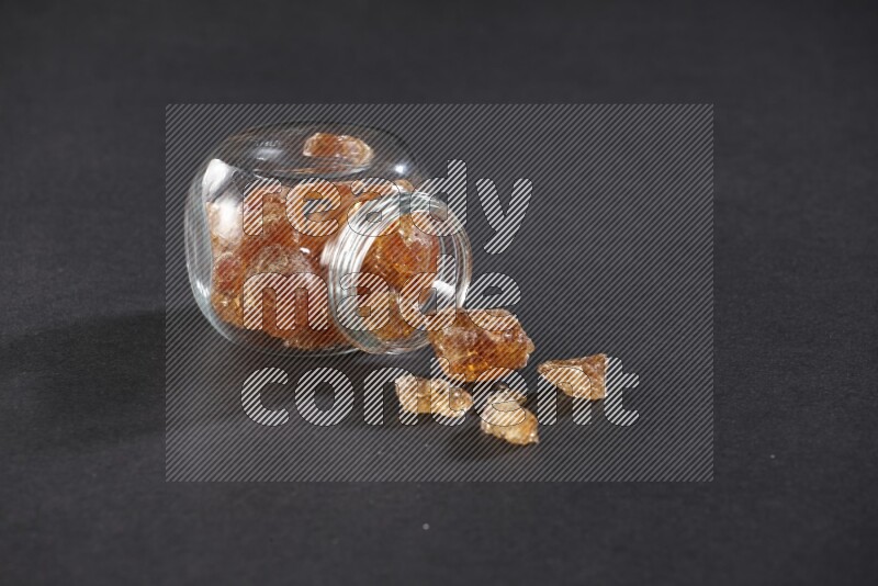 A glass spice jar full of gum arabic and jar is flipped with fallen pieces on black flooring