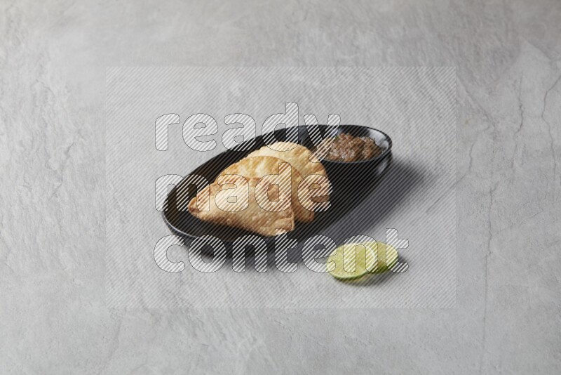 Three fried sambosas in an oval shaped black plate and a sauce in a black round ramekin with two lemon slices on the side on a gray background