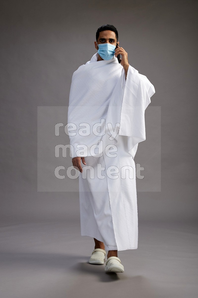 A man wearing Ehram with face mask Standing talking on phone on gray background