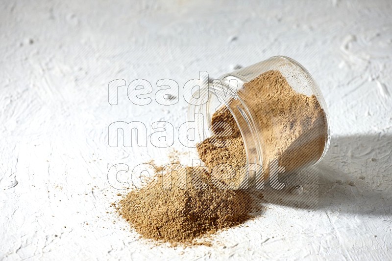 A flipped glass jar full of cumin powder and powder spilled out on textured white flooring