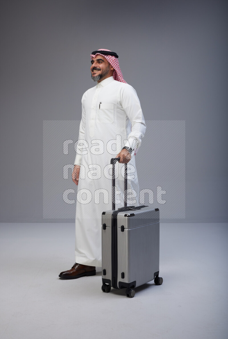Saudi man wearing Thob and red Shomag standing holding Travel bag on Gray background