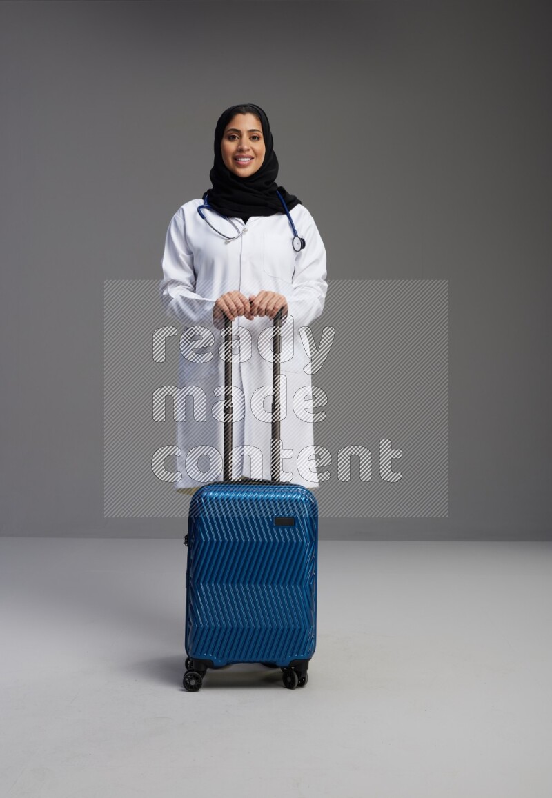 Saudi woman wearing lab coat with stethoscope standing holding Travel bag on Gray background