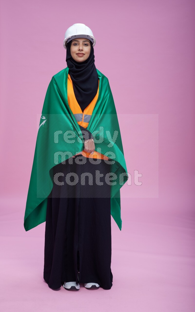 Saudi woman wearing Abaya with engineer vest and helmet standing holding Saudi flag on pink background