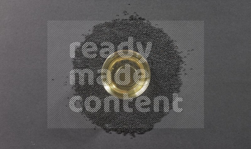 A glass bowl full of black seeds oil surrounded by the seeds on a black flooring