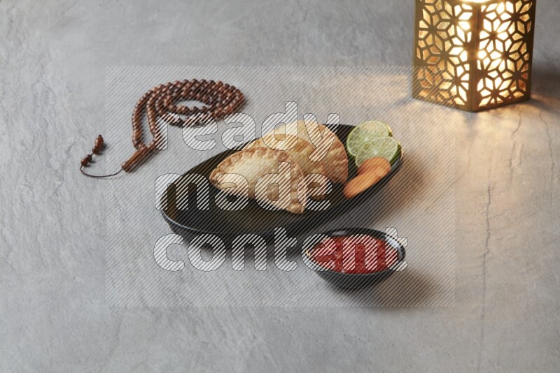 Three fried sambosas in an oval shaped black plate and a red sauce in a black round ramekin with a brown misbaha and a golden lantern on the side on a gray background