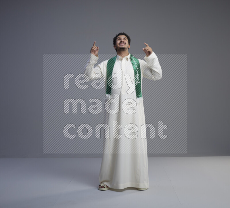 A Saudi man standing wearing thob and Saudi flag scarf on gray background