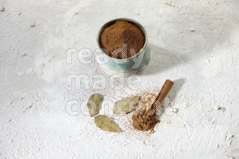 Cinnamon powder in a ceramic bowl with cinnamon sticks and laurel leaves on white background