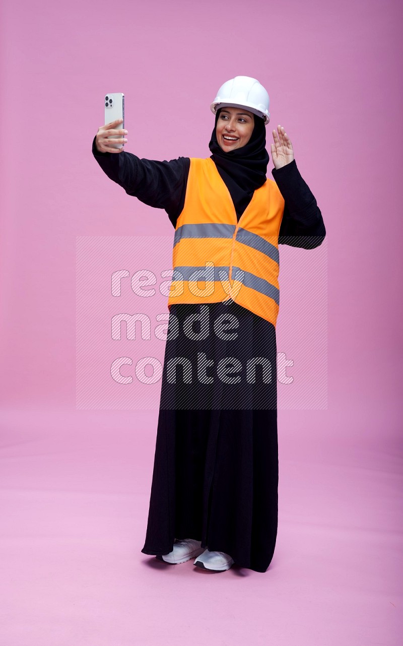 Saudi woman wearing Abaya with engineer vest and helmet standing taking selfie on pink background