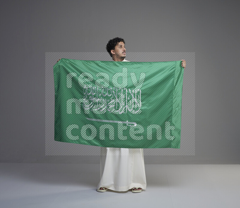 A saudi man standing wearing thob holding big saudi flag on gray background