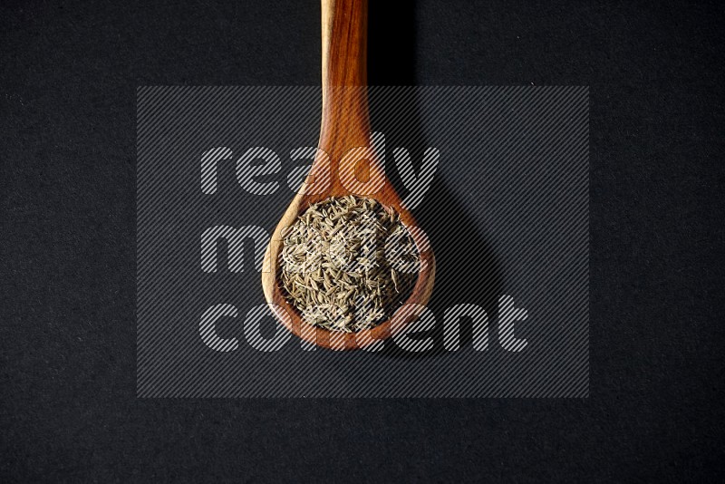 A wooden ladle full of cumin seeds on black flooring