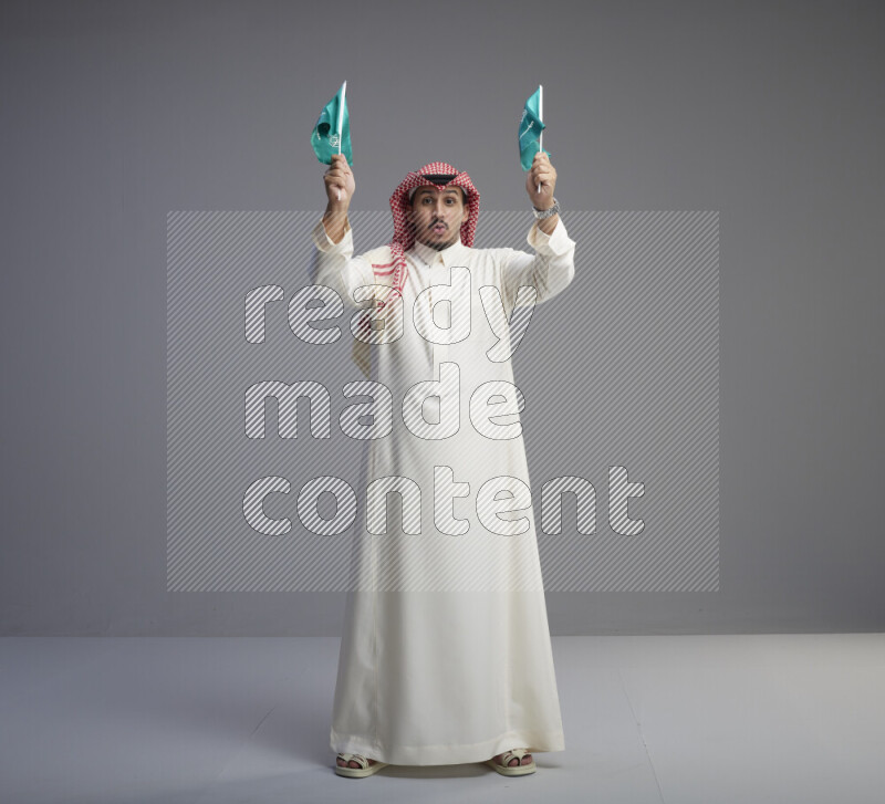 A Saudi man standing wearing thob and red shomag raising small Saudi flag on gray background