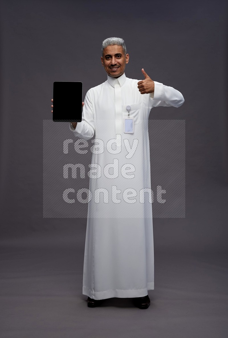 Saudi man wearing thob with pocket employee badge standing showing tablet to camera on gray background