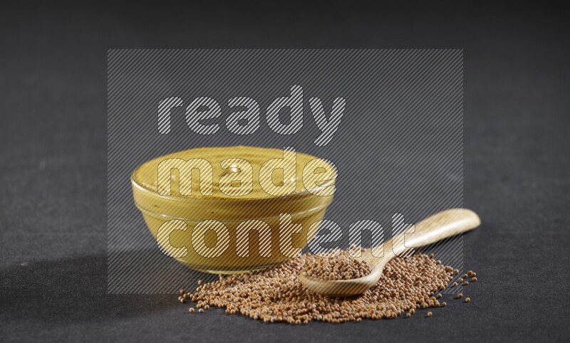 A glass bowl full of mustard paste with mustard seeds underneath and a full wooden spoon on black flooring