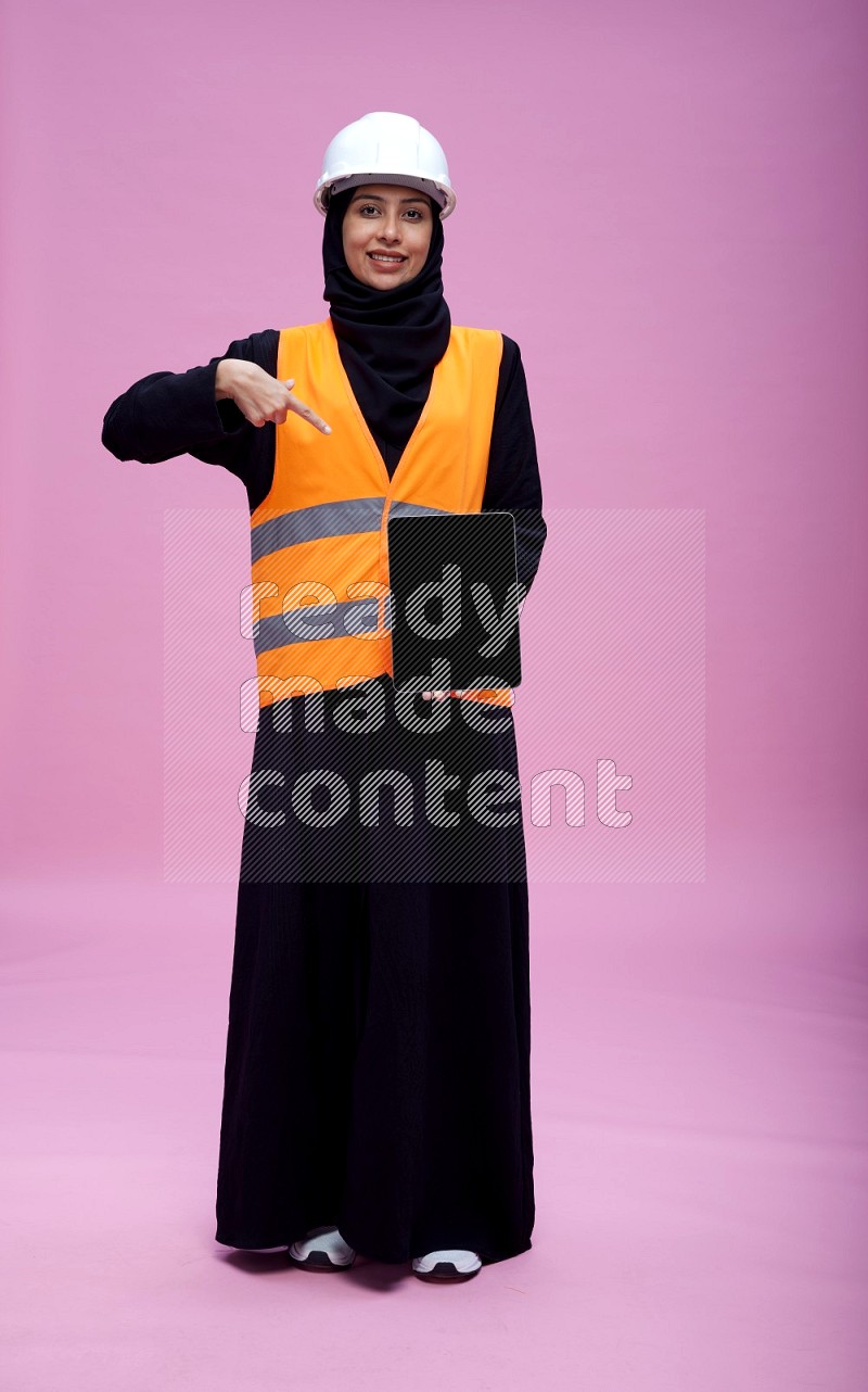 Saudi woman wearing Abaya with engineer vest and helmet standing showing tablet to camera on pink background