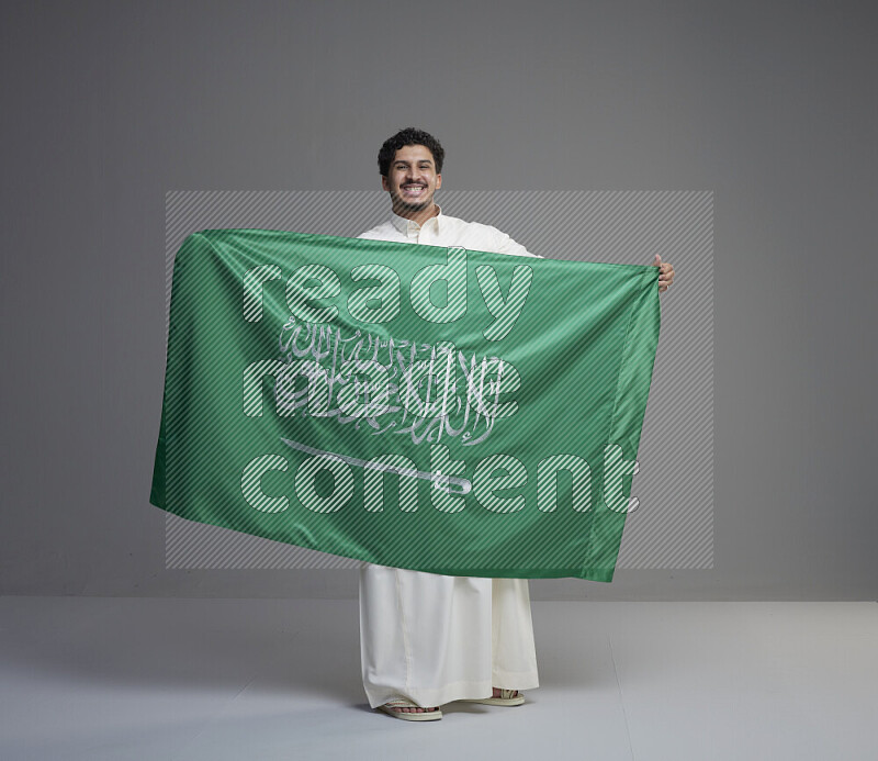 A saudi man standing wearing thob holding big saudi flag on gray background