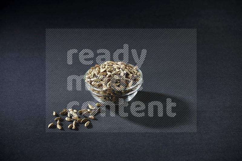 A glass bowl full of cardamom and more seeds spreaded beneath the bowl on black flooring