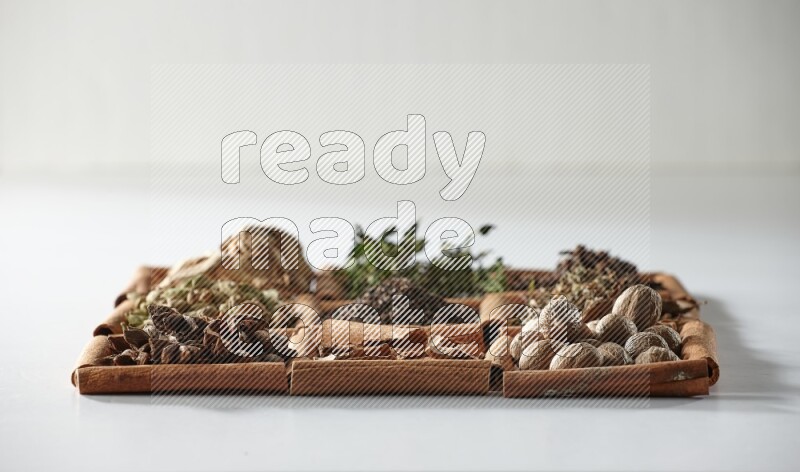 9 مربعات من أعواد القرفة ممتلئة بالشاي في المنتصف محاطة بجوزة الطيب والنعناع المجفف والقرنفل والريحان المجفف والزنجبيل المجفف والقرفة واليانسون والهيل على أرضية بيضاء