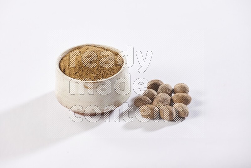 A beige pottery bowl full of nutmeg powder with whole seeds beside it on a white flooring