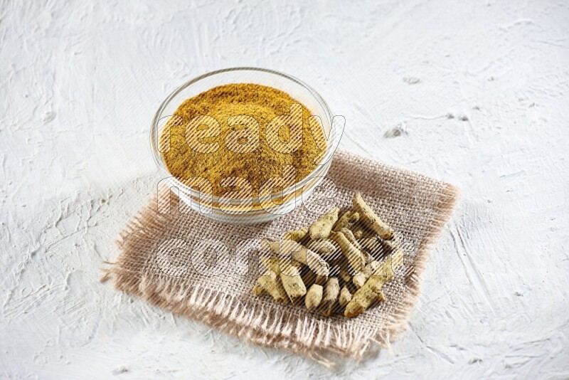 A glass bowl full of turmeric powder and dried turmeric whole finger on a piece of burlap on a textured white flooring