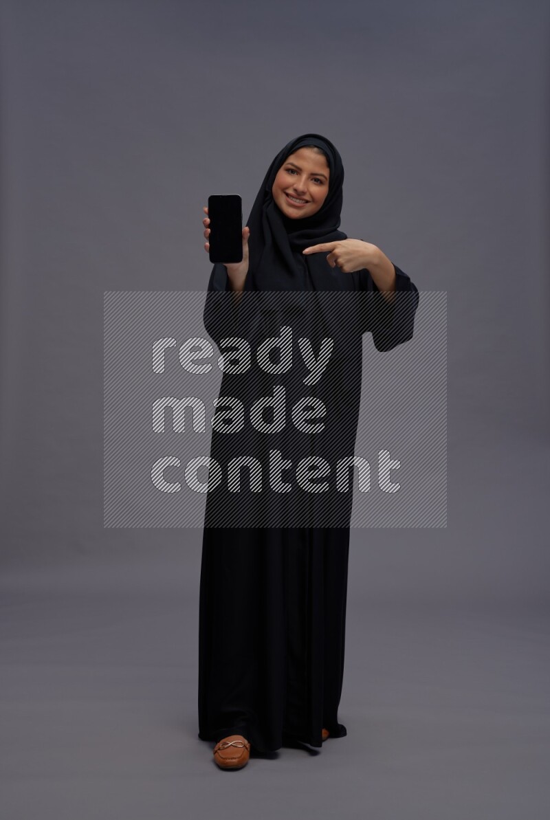 Saudi woman wearing Abaya standing showing phone to camera on gray background