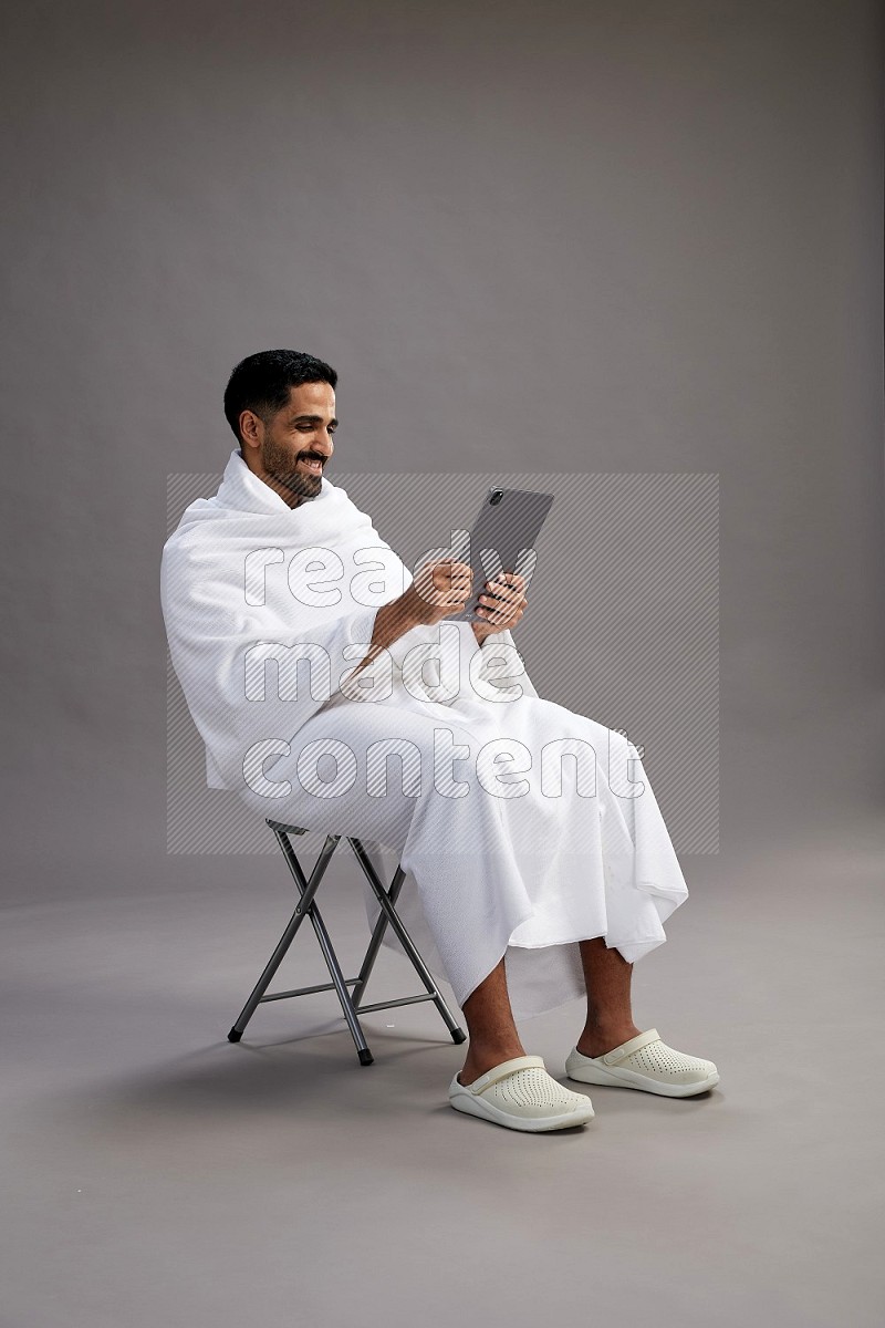 A man wearing Ehram sitting on chair holding a tablet on gray background