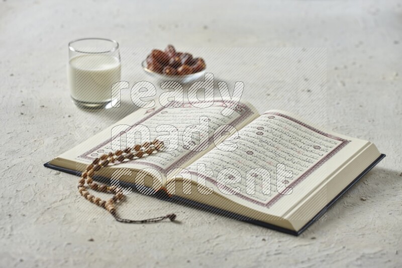 Quran with dates, prayer beads and different drinks all placed on textured white background