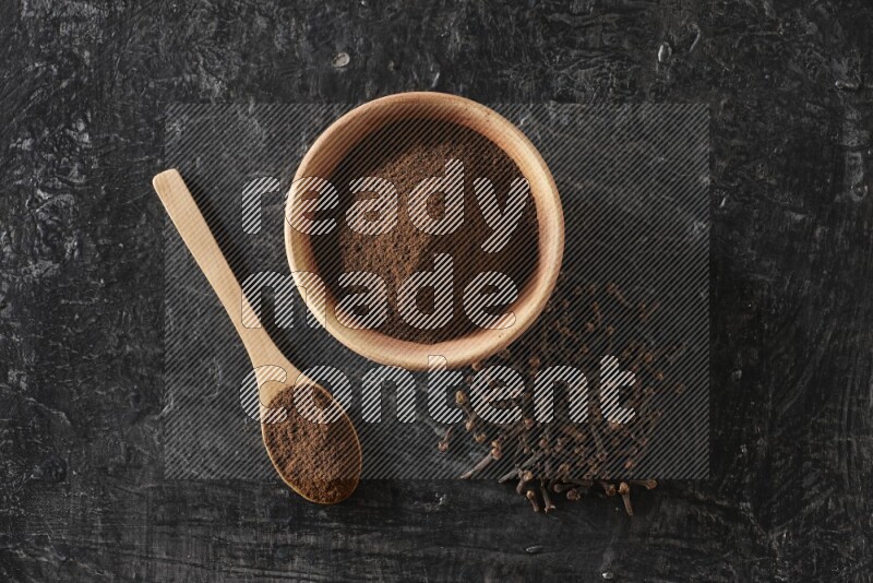 A wooden bowl and a wooden spoon full of cloves powder with spreaded cloves on a textured black flooring
