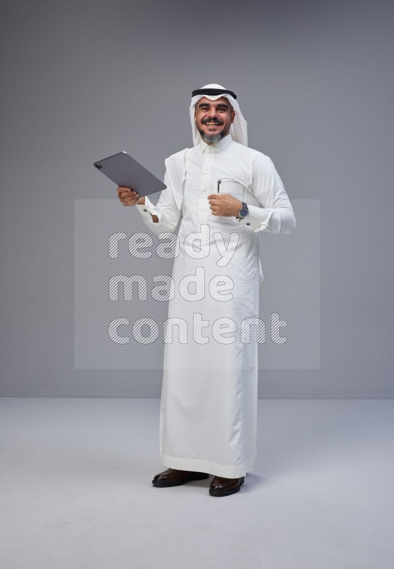 Saudi man Wearing Thob and white Shomag standing working on tablet on Gray background