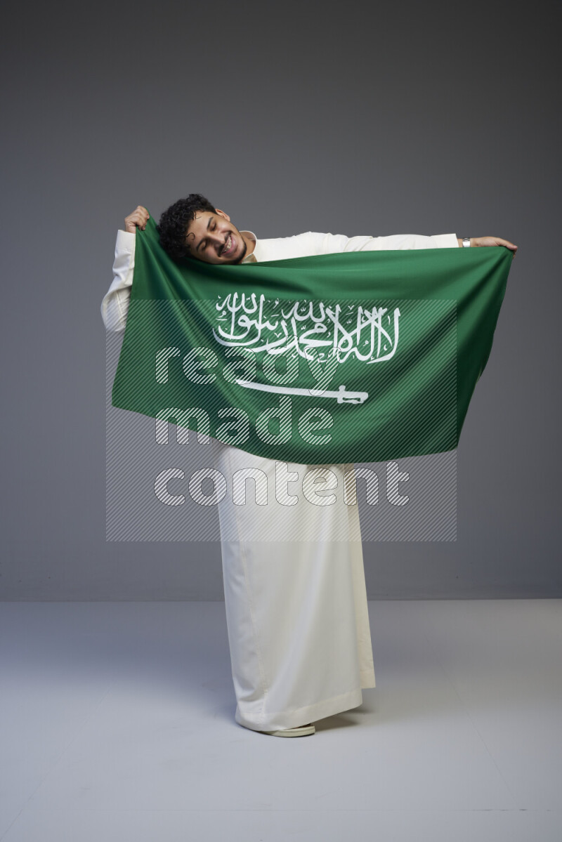 A Saudi man standing wearing thob holding big Saudi flag on gray background