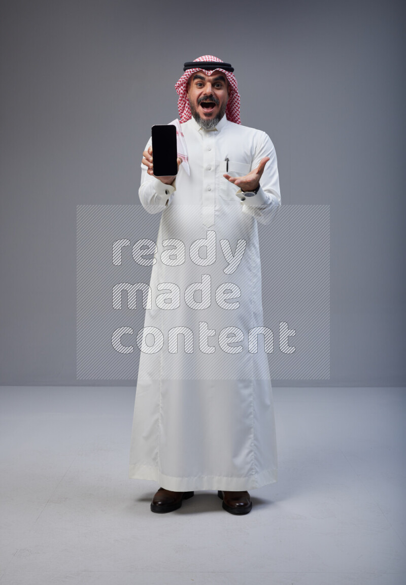 Saudi man Wearing Thob and red Shomag standing showing phone to camera on Gray background